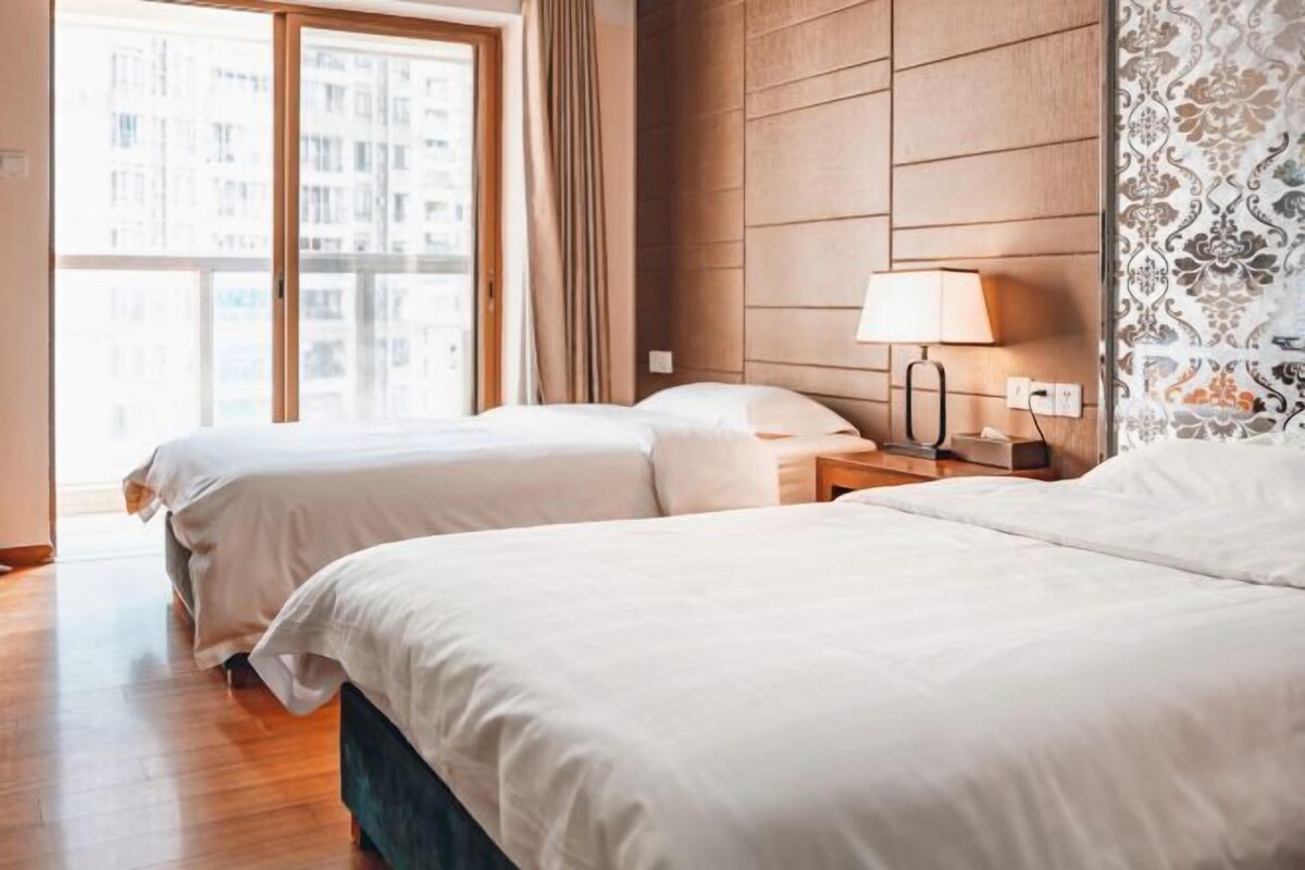 A hotel room with two beds, a wooden headboard, a nightstand with a lamp, and a window with curtains.