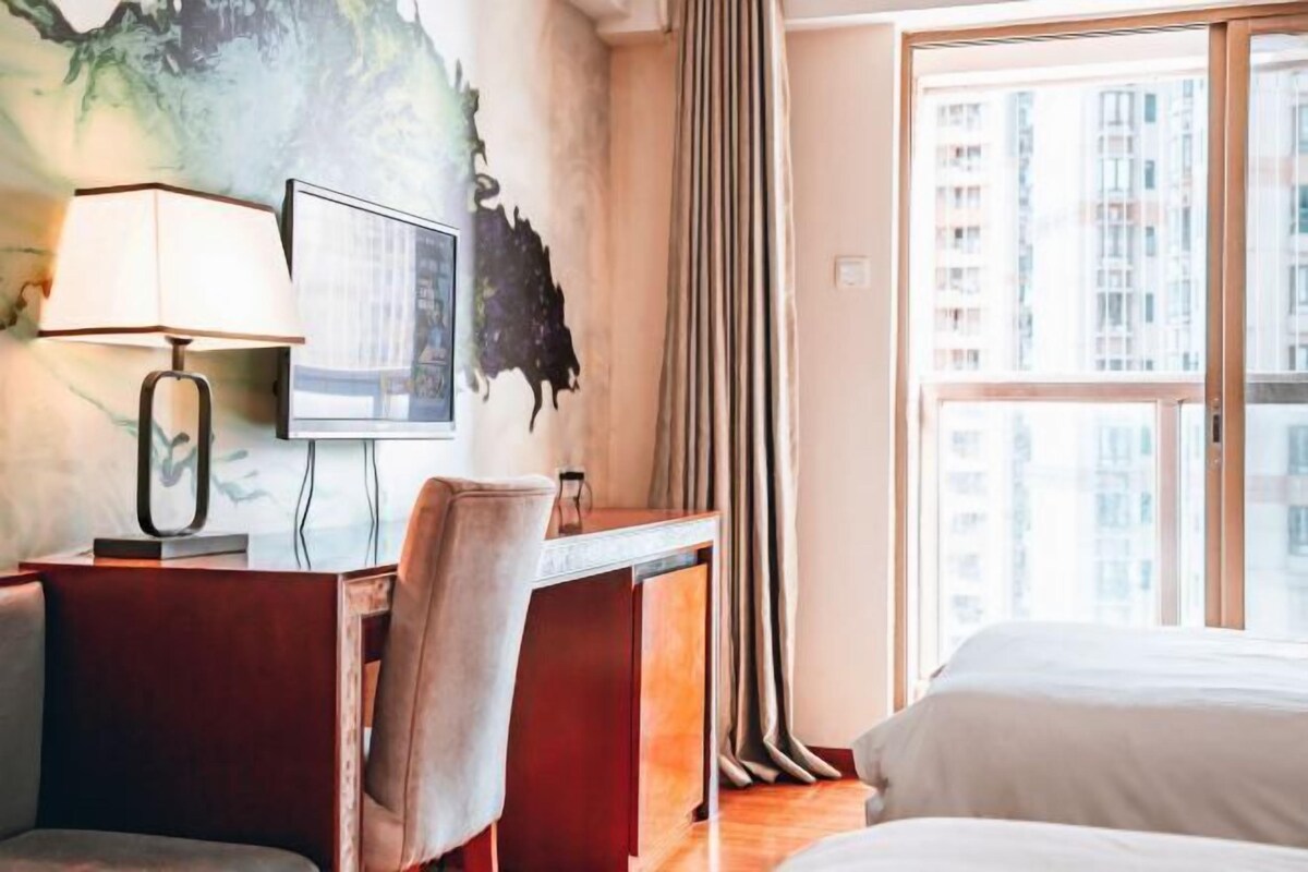 A hotel room with a desk, chair, bed, and a large window with a view of high-rise buildings.