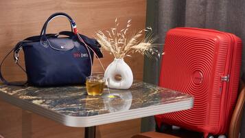 A table with a blue bag, a vase with dried plants, a glass of tea, and a red suitcase.