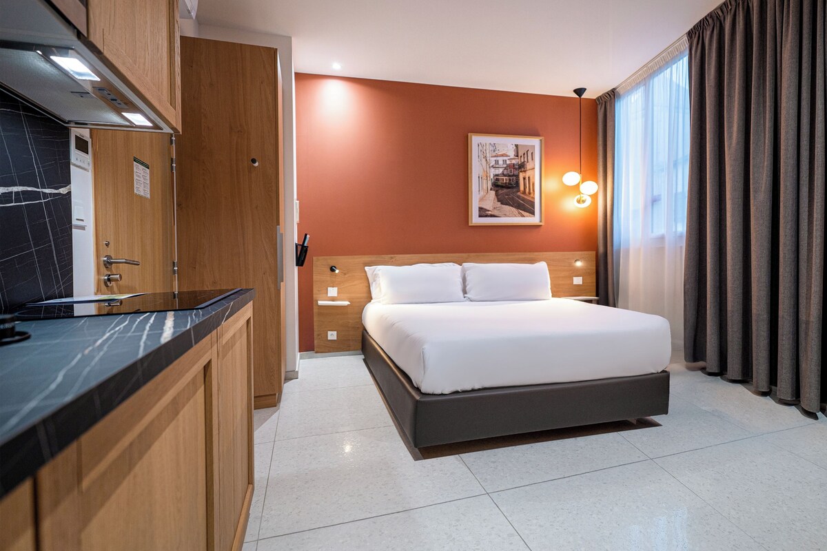 A modern hotel room with a bed, a small desk, a mirror, and a picture on the wall.