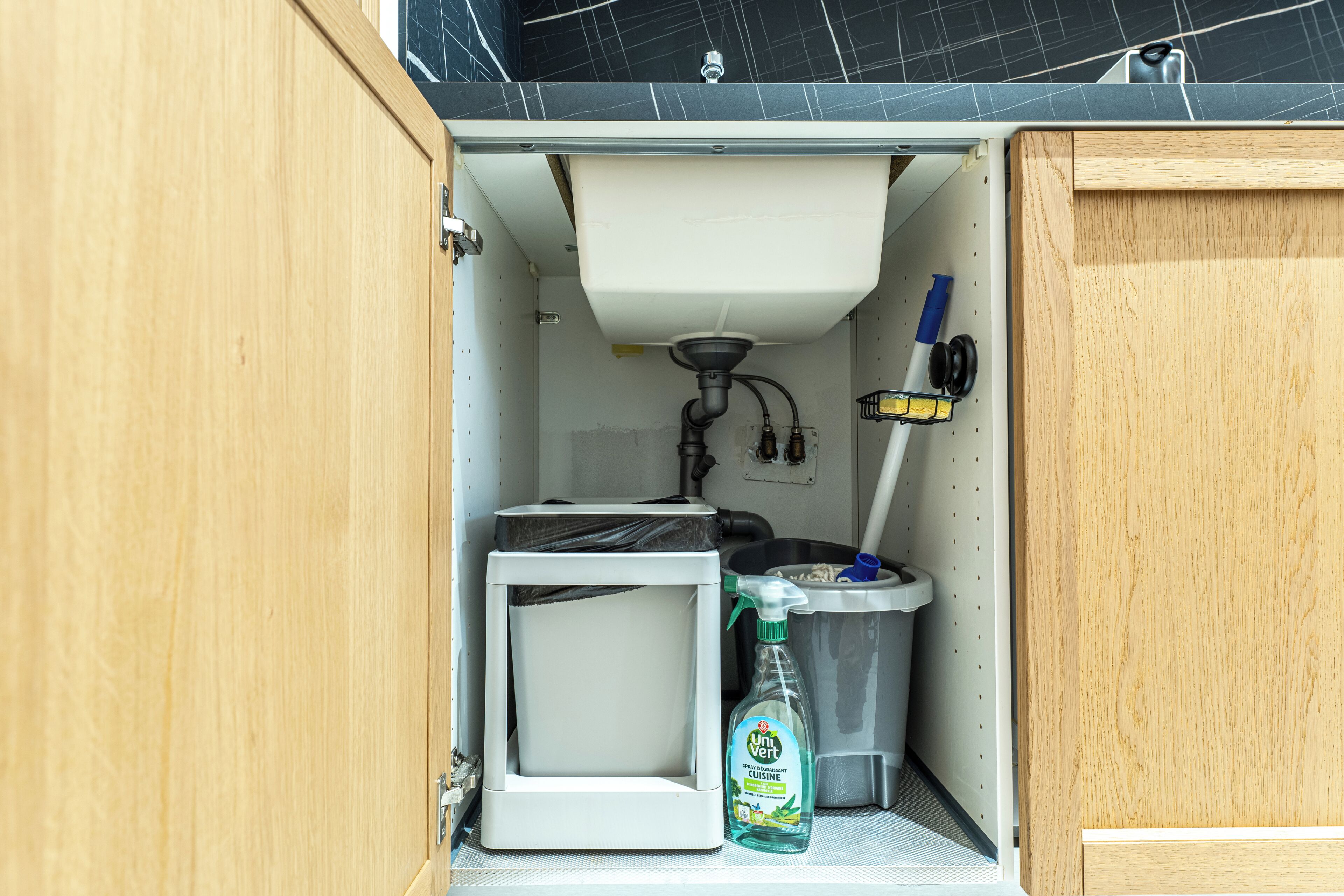 Una kitchenette compacta con un fregadero, un refrigerador pequeño, un cesto de basura y una botella de limpiador.