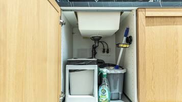 Una kitchenette compacta con un fregadero, un refrigerador pequeño, un cesto de basura y una botella de limpiador.