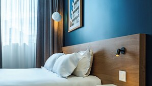 A hotel room with a bed, a nightstand, a framed picture on the wall, and a window with curtains. - Corner Set (Cholet)