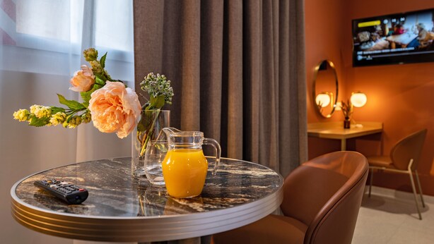 A hotel room with a round table, a remote control, a vase with flowers, and a glass pitcher of orange juice. - Corner Set (Cholet)