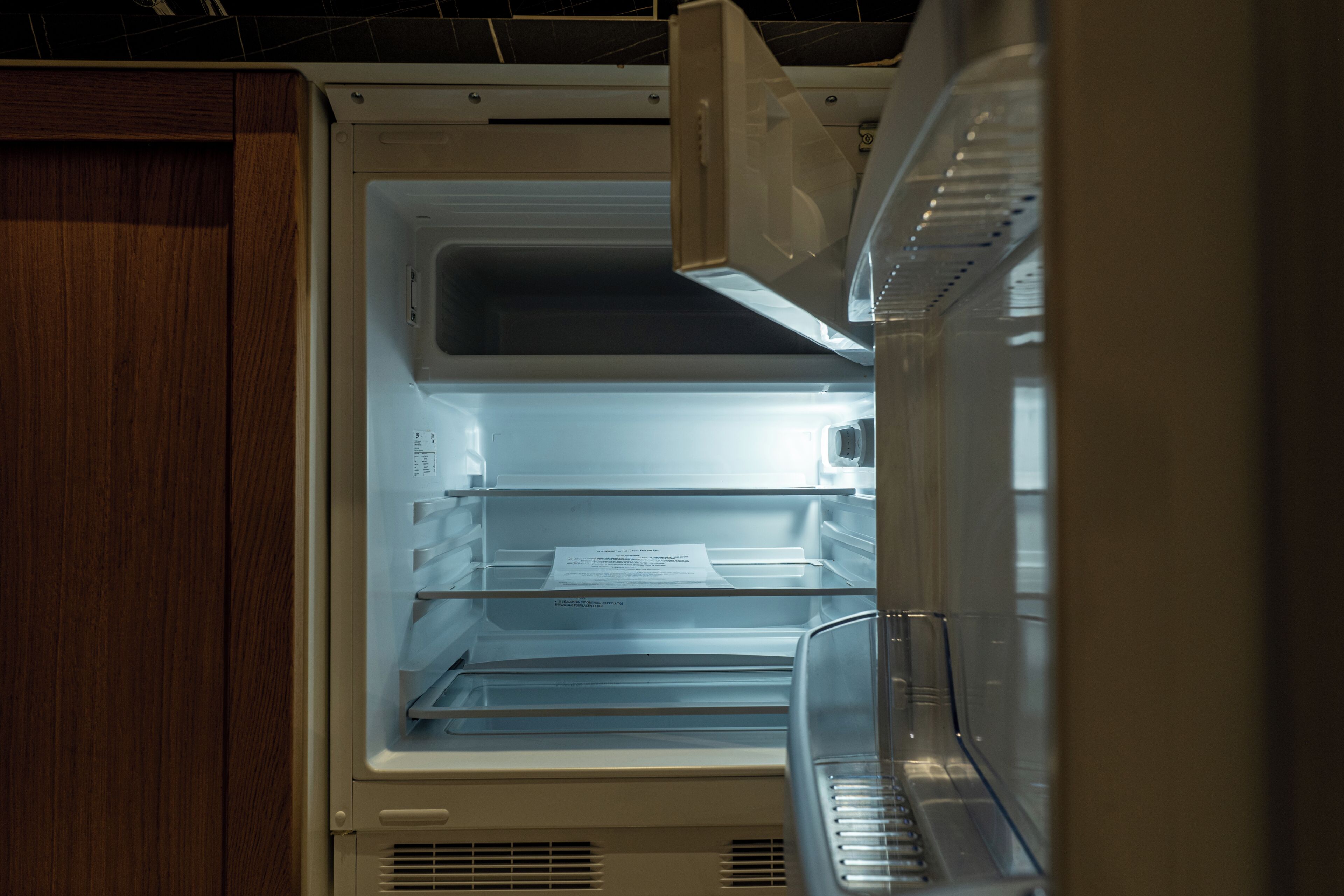 An open refrigerator with a paper inside, a wooden cabinet door, and a vent at the bottom.