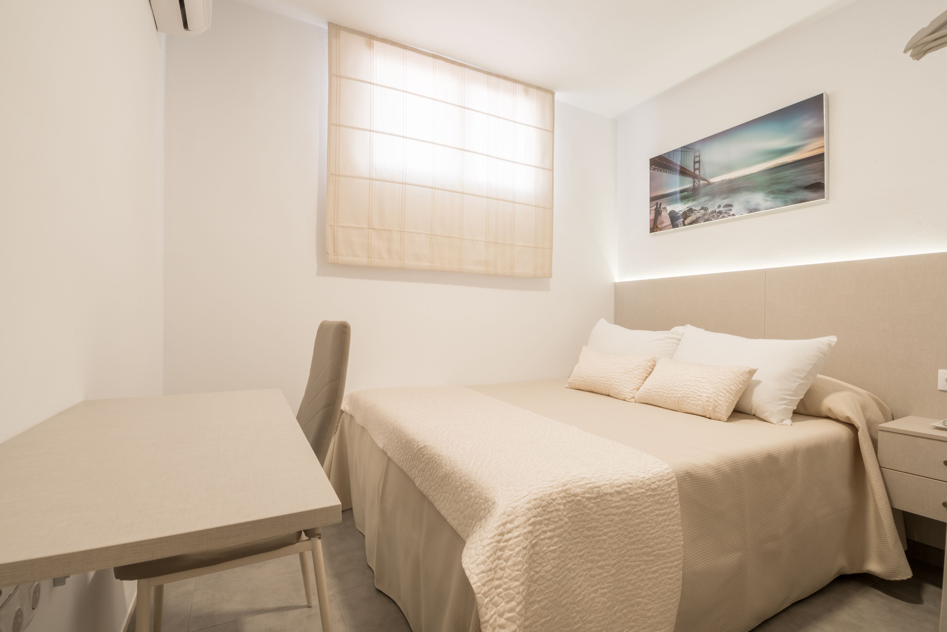 A hotel room with a bed, a desk, a chair, a window with blinds, and a painting on the wall.