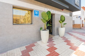 A modern building with a sign reading "HOSTAL" and a blue sign with "H" and "CIUDAD". There are two large potted plants in front.