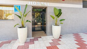 A hotel entrance with the sign "HOSTAL" above the door, large potted plants on either side, and a tiled walkway leading to the entrance.