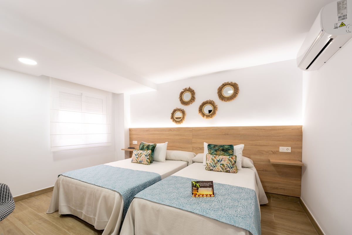 A hotel room with two beds, a wooden headboard, a window with blinds, and wall-mounted air conditioning.