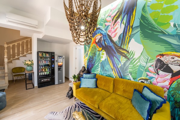 A living room with a yellow sofa, a colorful mural, a vending machine, and a staircase.