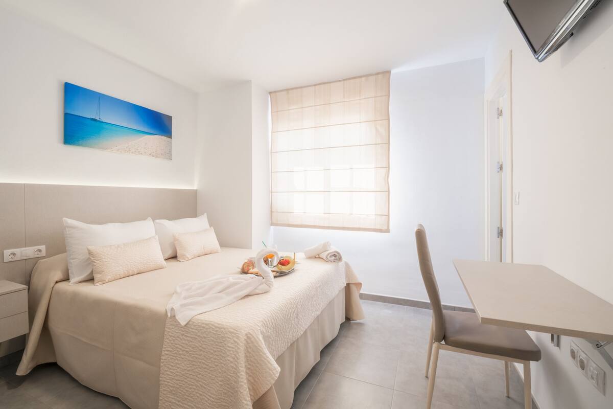 A hotel room with a bed, a dining table, a chair, a window with blinds, and a wall-mounted TV.