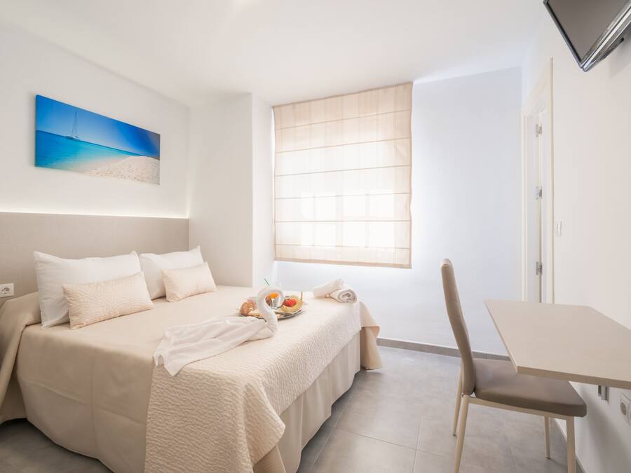 Een hotelkamer met een bed, een eettafel, een stoel, een raam met jaloezieën en een aan de muur bevestigde televisie.