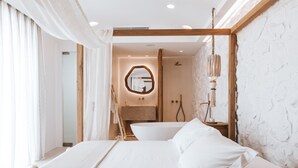 A modern bedroom with a large bed, a round mirror, a bathtub, and a wooden ceiling beam. - Cocopalm Villas Naxos (Naxos)
