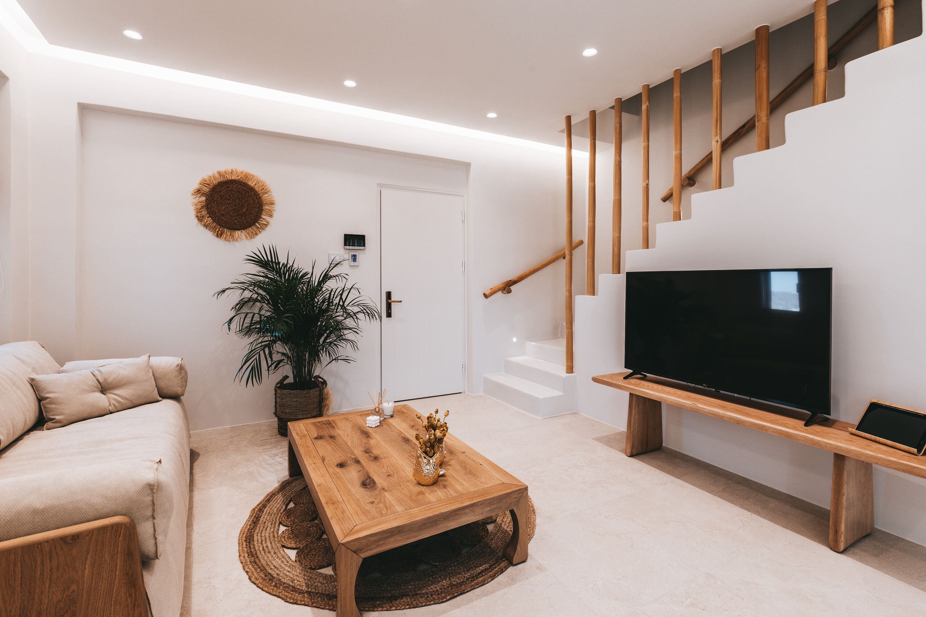 A modern living room with a sofa, a wooden coffee table, a TV on a wooden stand, and a staircase with bamboo railings.