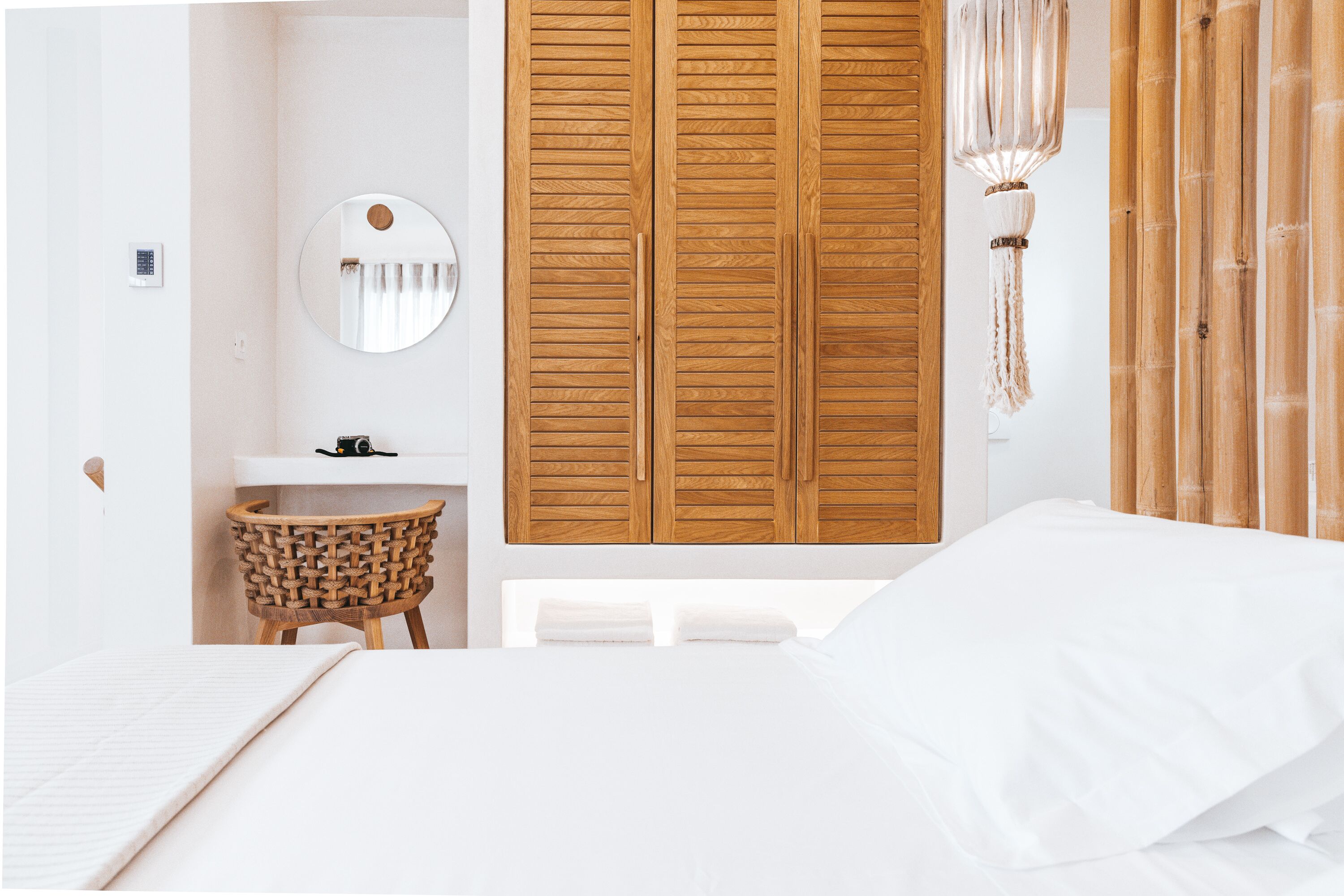 Een slaapkamer met een bed, een ronde spiegel, een houten garderobe, een klein rond tafeltje en een hangende lamp.