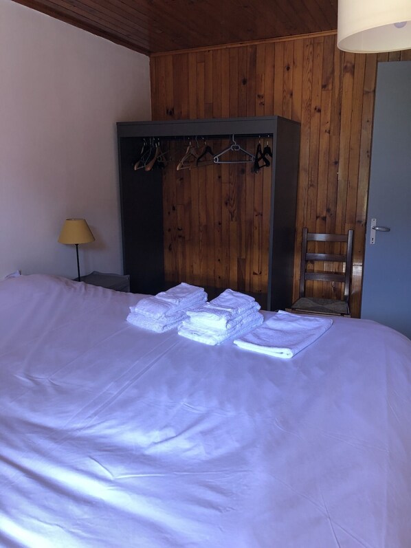 Een net opgemaakt bed met opgevouwen handdoeken, een houten garderobe met kledinghangers, een stoel en een lamp in een kamer met houten lambrisering.