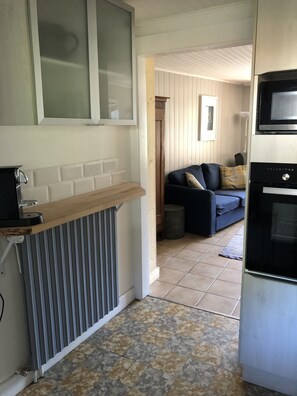 A compact kitchen with a built-in oven, a small countertop, and a radiator. A dark-colored sofa is visible in the background, and the floor is tiled.