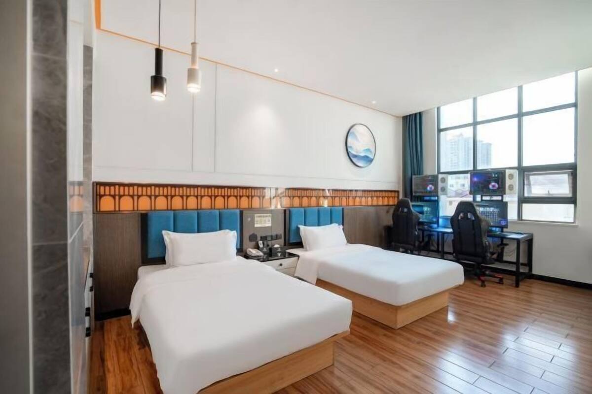 Une chambre d’hôtel avec deux lits, un bureau avec des ordinateurs, une horloge au mur et une fenêtre donnant sur la ville.