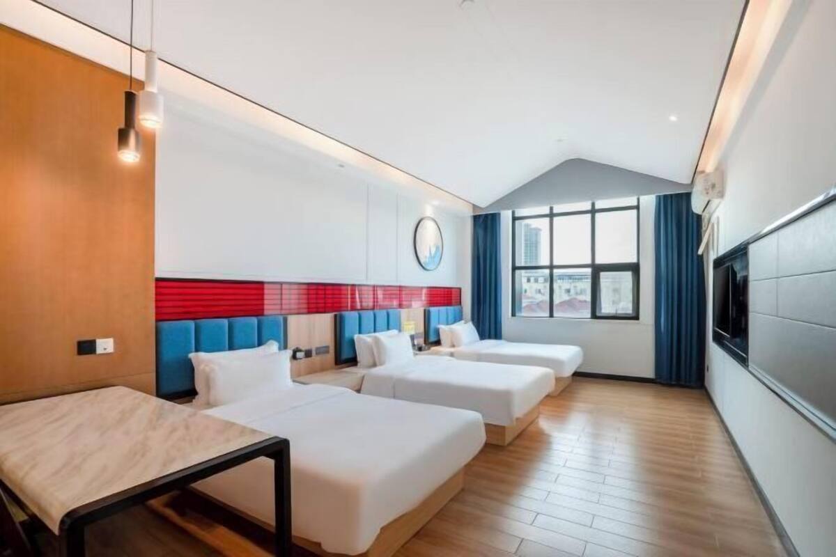 Une chambre d’hôtel avec trois lits, un bureau, une grande fenêtre avec des rideaux et une vue sur la ville.