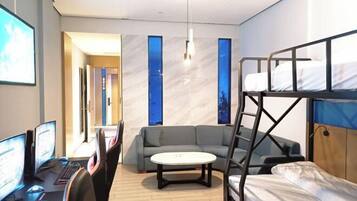 A modern hotel room with a bunk bed, a sofa, a desk with computers, and a large window.