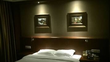 A hotel room with a bed, two framed pictures on the wall, a nightstand with a lamp, and a curtain on the left.