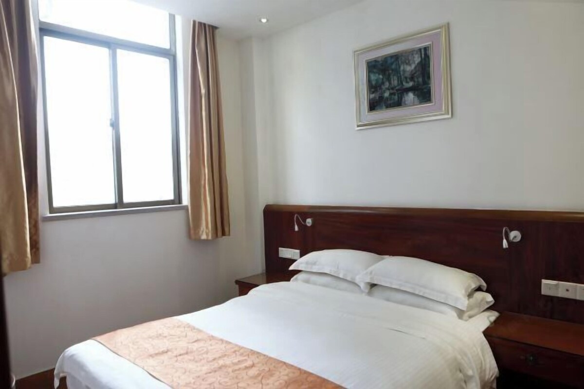 A hotel room with a bed, a nightstand, a window with curtains, and a painting on the wall.