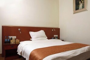 Habitación de hotel con cabecera de madera, cama con ropa de cama blanca, manta marrón, mesita de noche con lámpara y un cuadro en la pared.