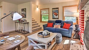 A cozy living room with a sofa, coffee table, and a lamp. There are stairs leading to another level, and a window with a view of trees. - Steps to Lake: Cabin w/ Deck in Lake Arrowhead (Lake Arrowhead)