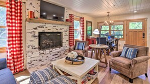A cozy living room with a fireplace, a television mounted above it, and a dining area with a table and chairs. - Steps to Lake: Cabin w/ Deck in Lake Arrowhead (Lake Arrowhead)