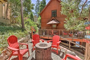 Une cabane en bois dotée d’une terrasse, entourée d’arbres et comprenant un foyer extérieur.