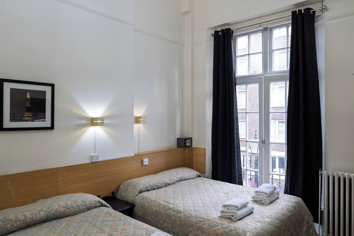 A hotel room with two beds, a framed picture on the wall, a nightstand, and a window with curtains.