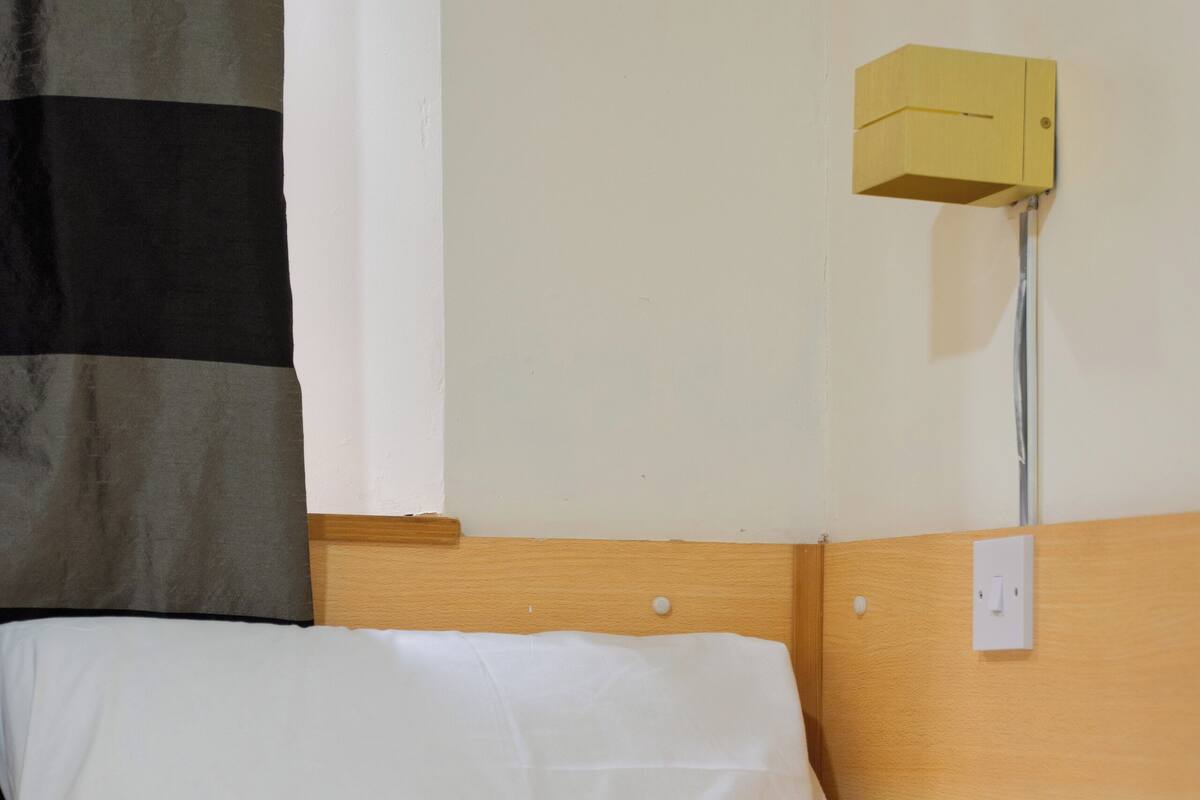 A hotel room with a bed, white bedding, a wooden headboard, a wall-mounted light fixture, and a power outlet.