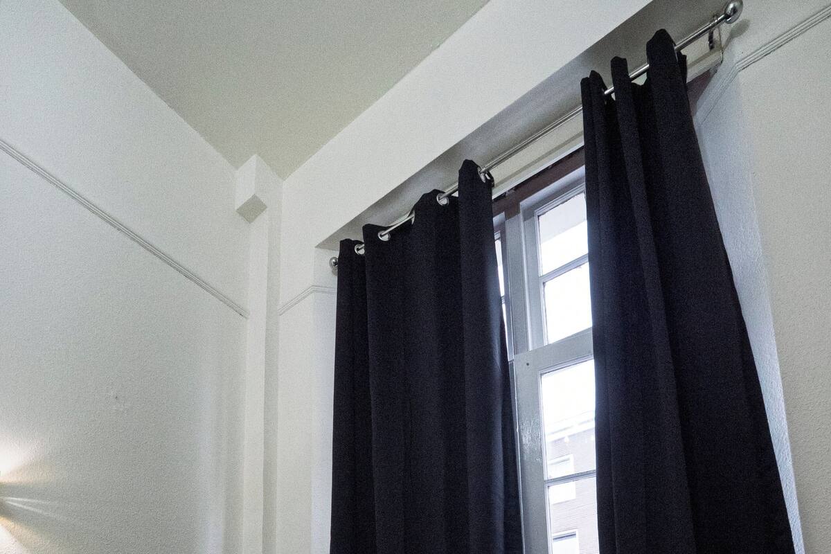 A hotel room with a bed, a bedside table with a clock, and a window with dark curtains.