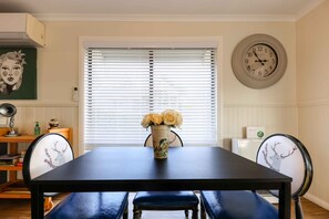 Un comedor con una mesa, sillas, un jarrón con flores, un reloj y una ventana con persianas.
