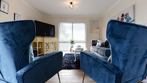 Un salon avec deux fauteuils bleus, un téléviseur, un canapé, une table basse et une fenêtre avec des stores.