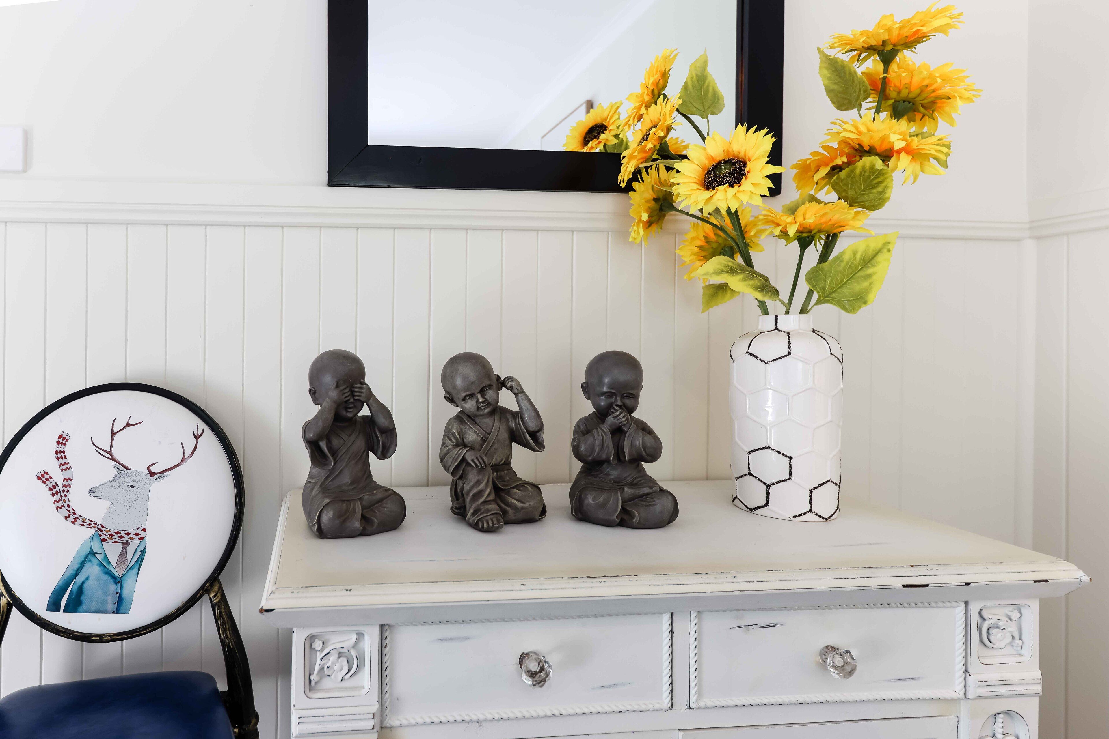 A white dresser with three bronze Buddha statues, a framed picture of a deer, and a vase with sunflowers.