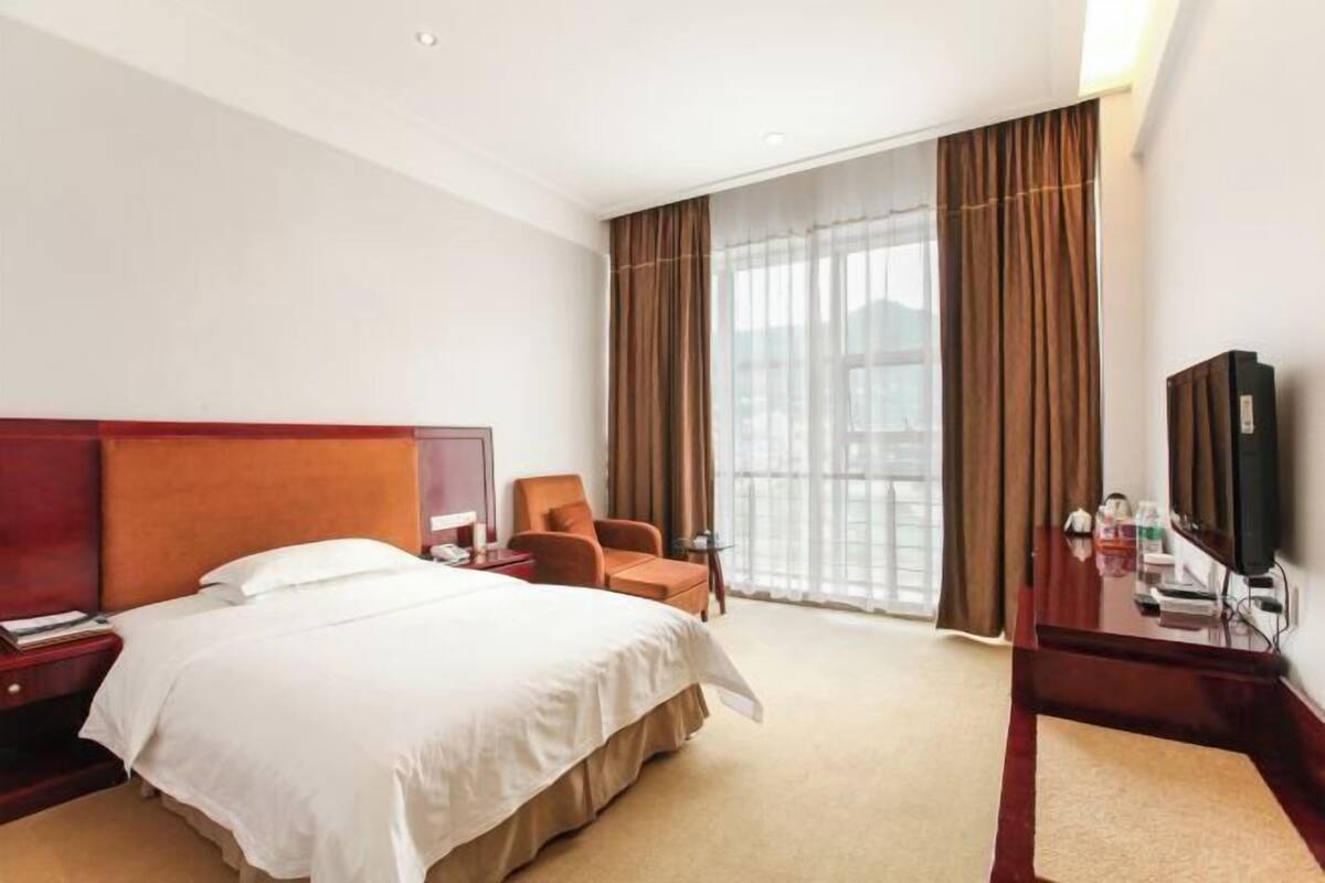 Quarto de hotel com uma cama grande, uma escrivaninha, uma cadeira, uma televisão e uma janela com cortinas.