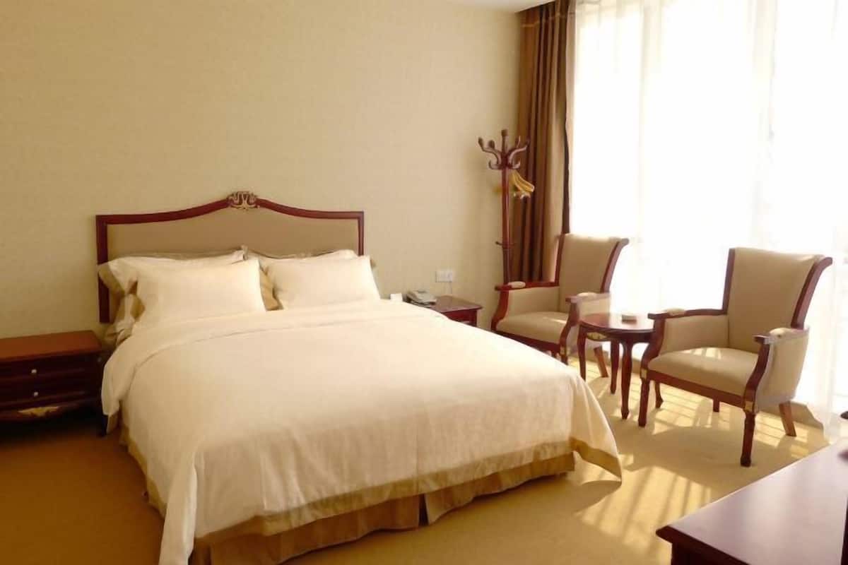 Quarto de hotel com uma cama grande, duas mesinhas de cabeceira, uma cadeira, uma mesa pequena e um cabideiro.