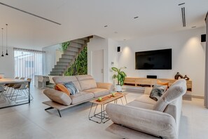 A modern living room with a sofa, coffee table, and TV. There is a staircase with greenery, a dining area, and a large window.