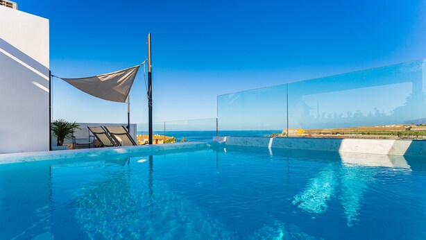 A clear blue pool with a view of the sea, a white building, and a shaded area with lounge chairs. - Green House - modern villa with a private rooftop pool in the south of Tenerife! (Guía de Isora)