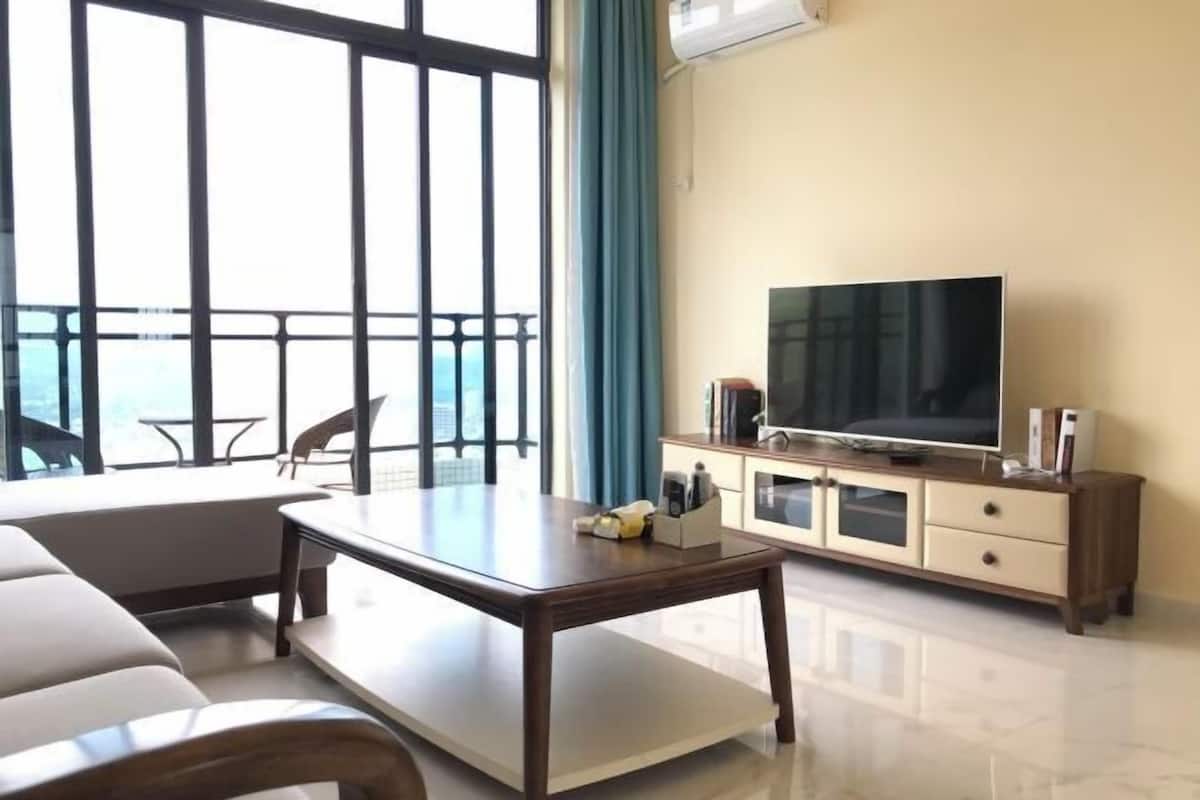 A modern living room with a sofa, a coffee table, a TV on a cabinet, and a balcony with a view.