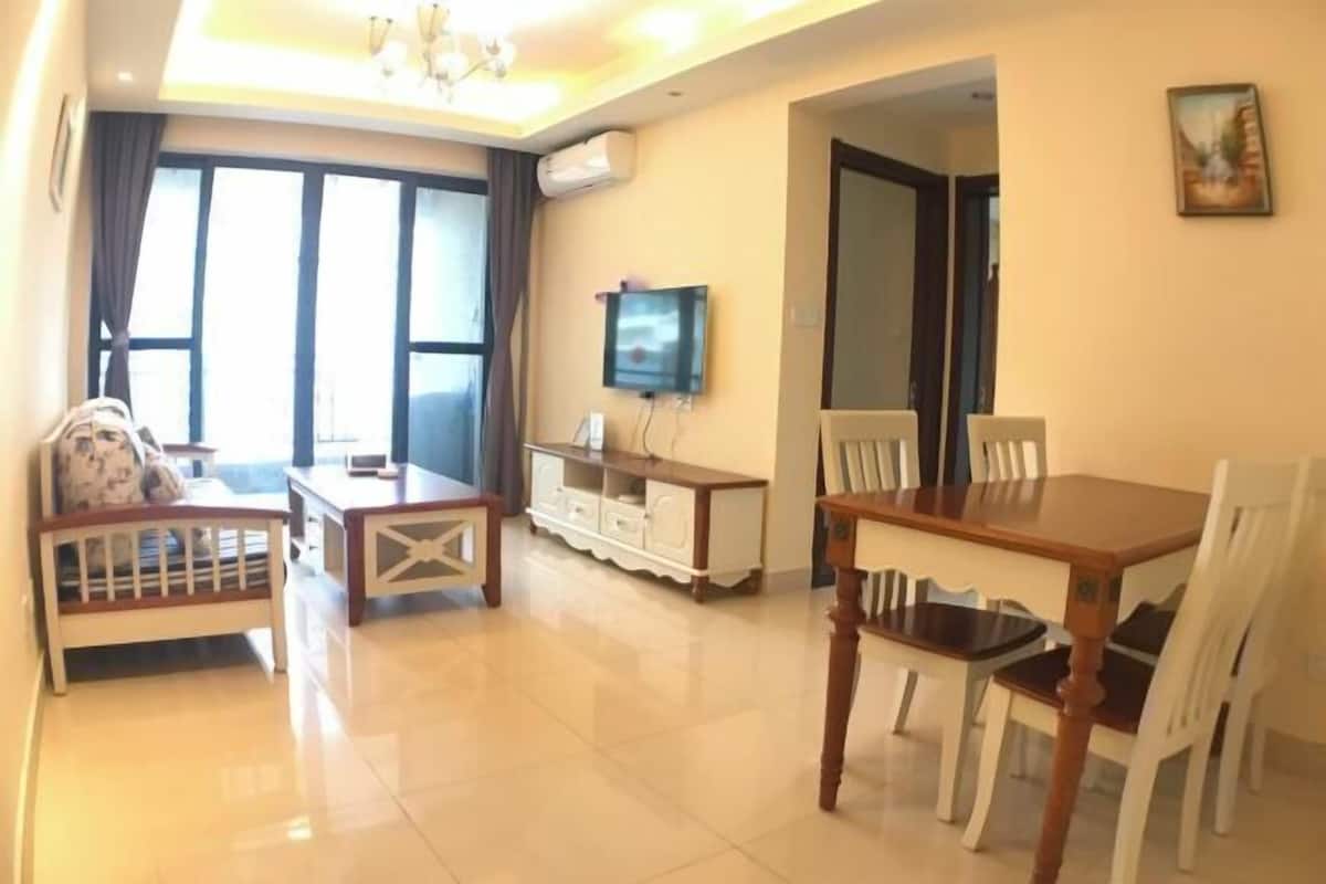 A spacious living area with a dining table and chairs, a sofa, a TV mounted on the wall, and a large window with curtains.
