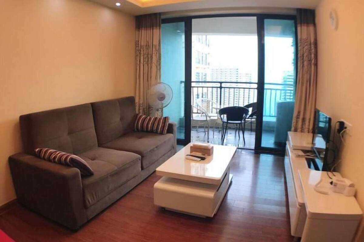 A living room with a sofa, a coffee table, a fan, a balcony with a chair, and a view of buildings.