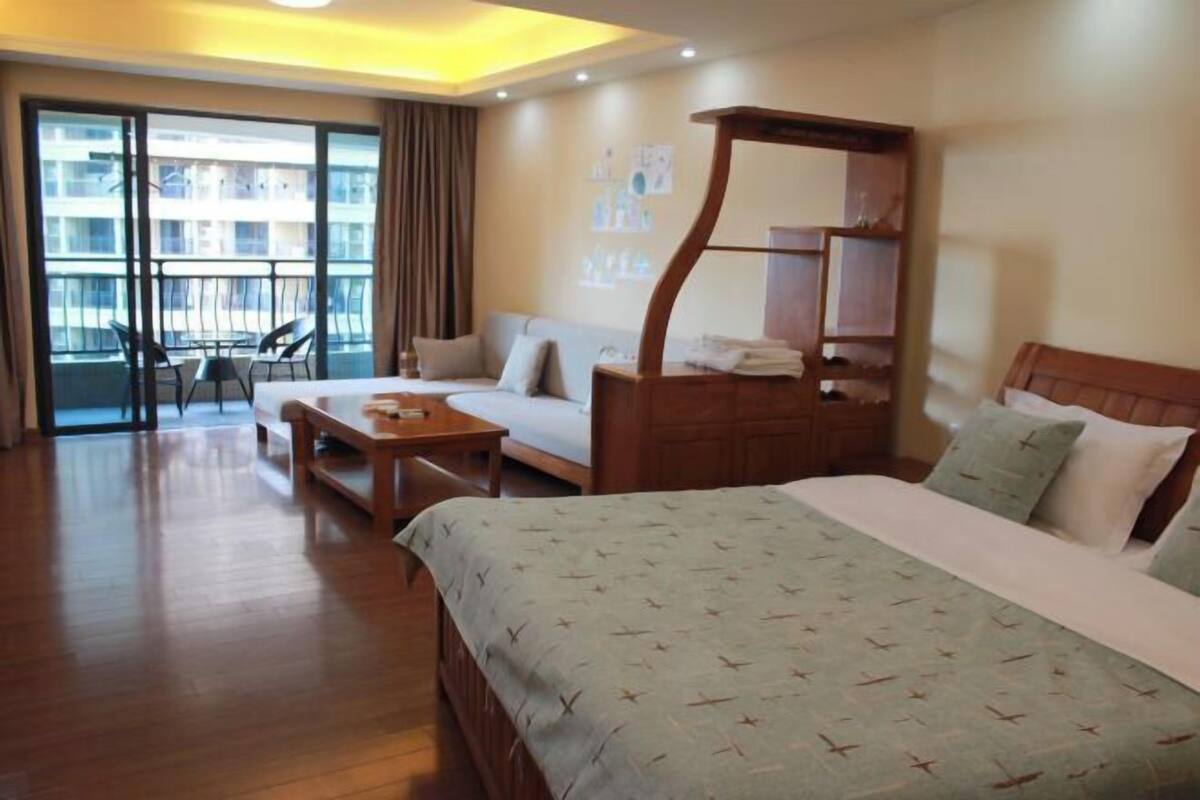 A hotel room with a bed, a desk, a chair, a balcony with a view of buildings, and a wooden nightstand.