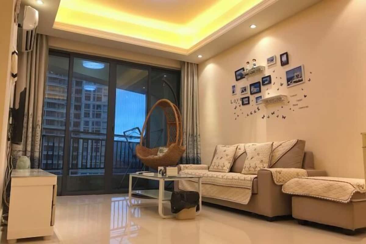 A modern living room with a sofa, a glass coffee table, and a hanging chair. There are pictures on the wall and a large window with a view of buildings.