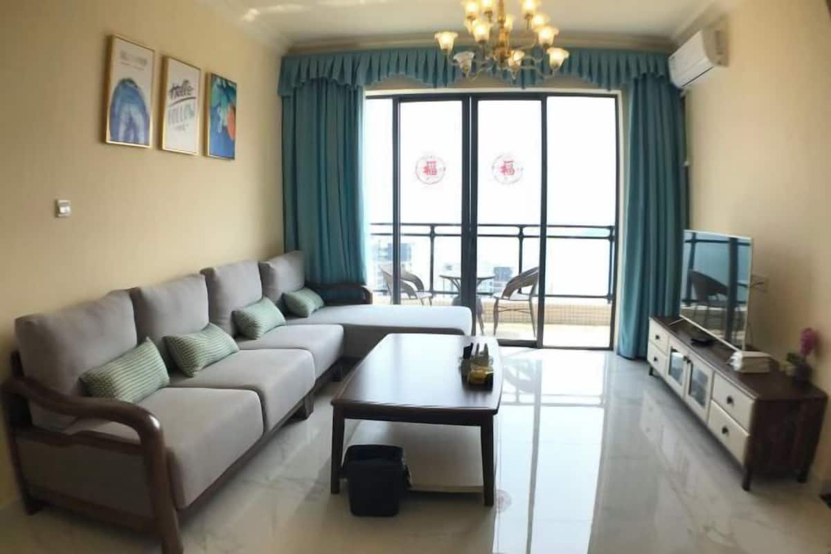 A living room with a sofa, coffee table, and television. There is a balcony with blue curtains and a chandelier.