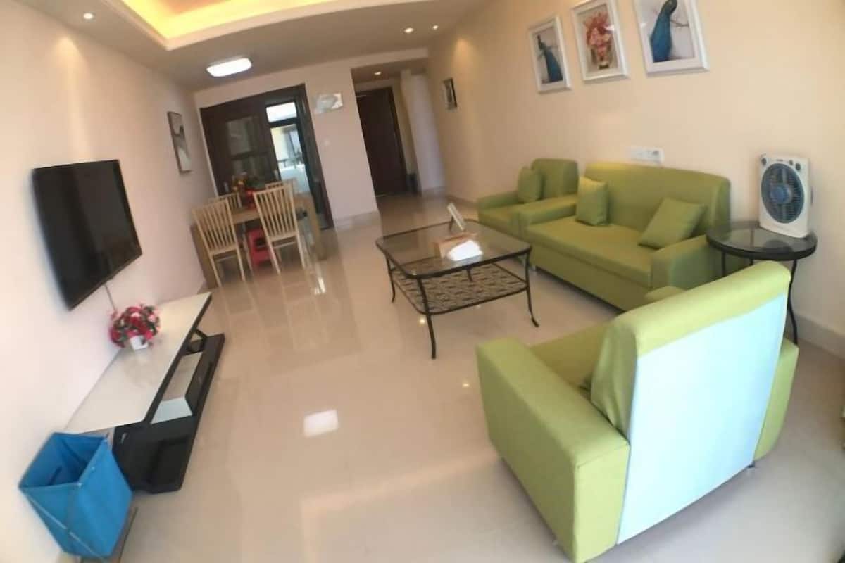 A modern living room with a green sofa, a glass coffee table, a flat-screen TV, and a fan.