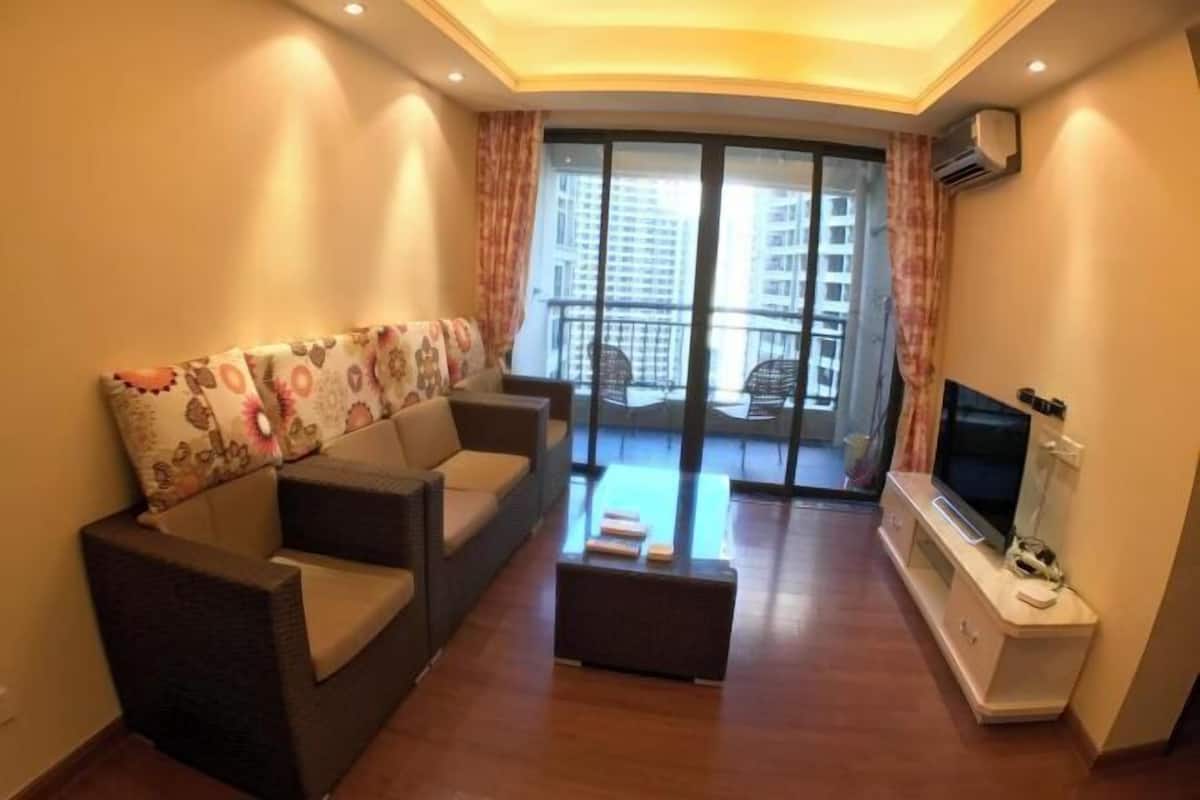 A living room with a sofa, a coffee table, a TV, and a balcony with a view of buildings.