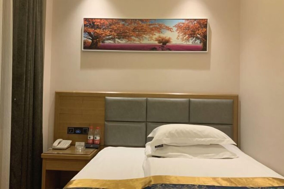 A hotel room with a bed, a nightstand with a phone and a bottle, and a painting of a tree on the wall.