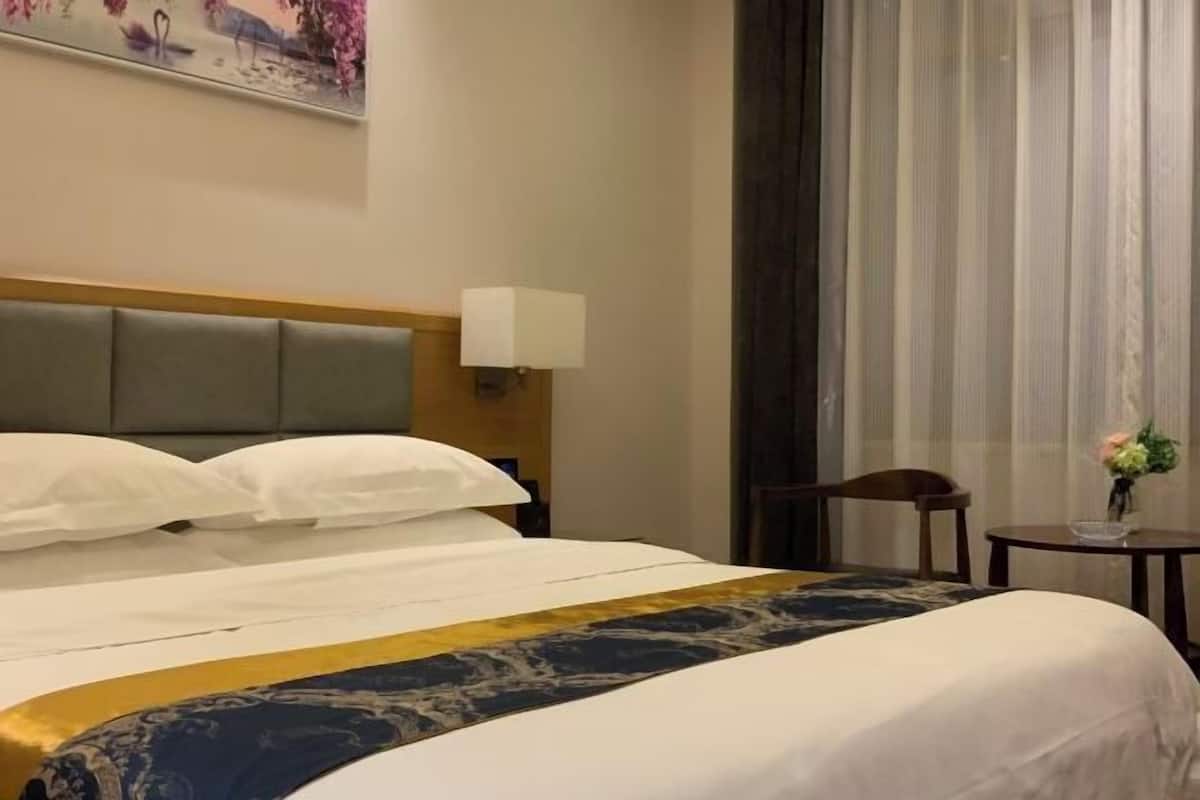 Habitación de hotel con una cama grande, cabecera, mesita de noche con lámpara, una silla y una pequeña mesa con un jarrón de flores.